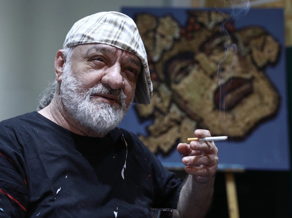 Saimir Strati holding a cigarette, with one of his mosaics behind him